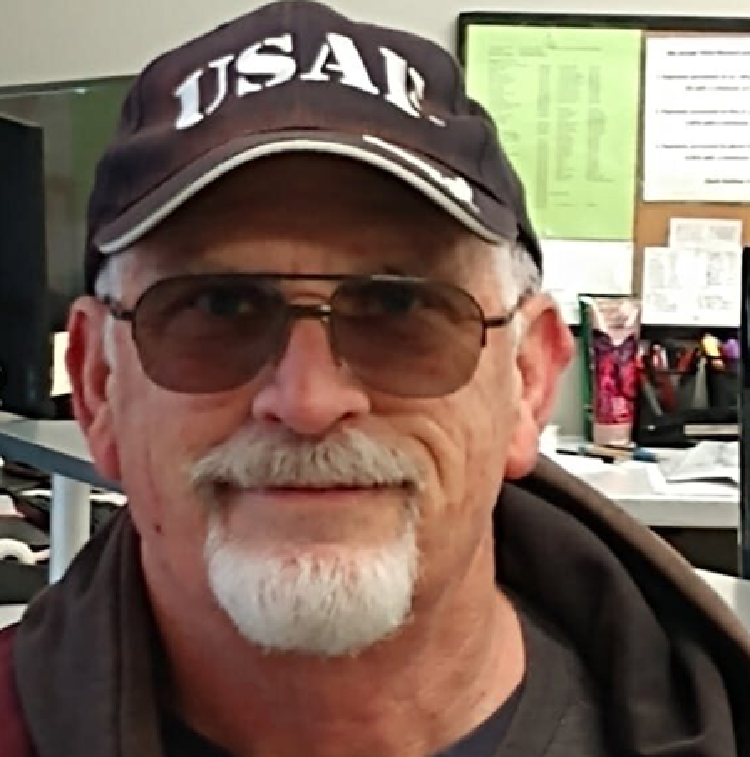 Older man with gray goatee and sunglasses wearing a dark cap with white lettering, in an office setting.